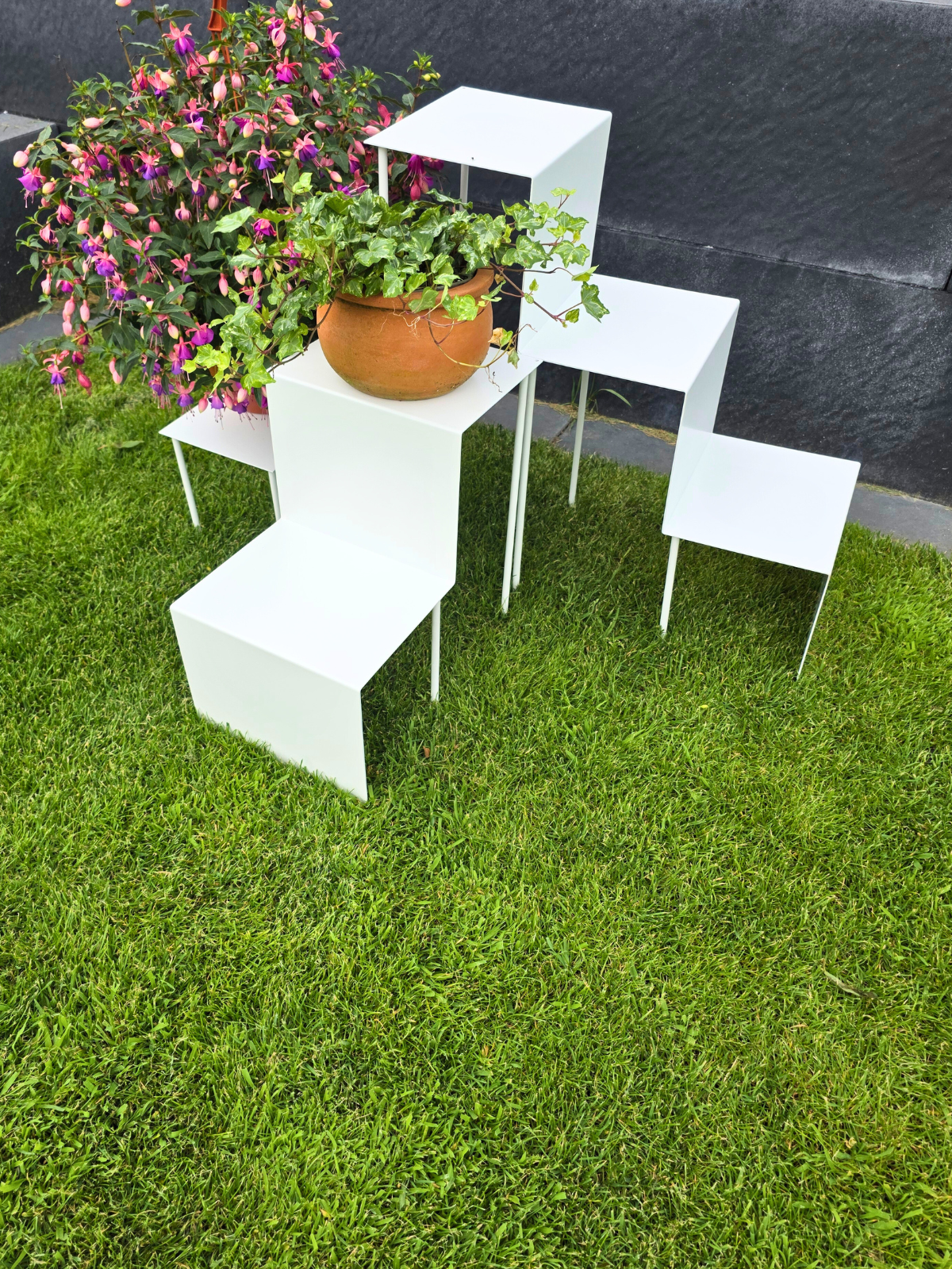 Zdjęcie przedstawia duży kwietnik składający się z trzech różnych wielkości części. Kwietnik jest cały w kwiatach w glinianych doniczkach. Kwietnik jest biały. Stoi na trawie w ogrodzie.