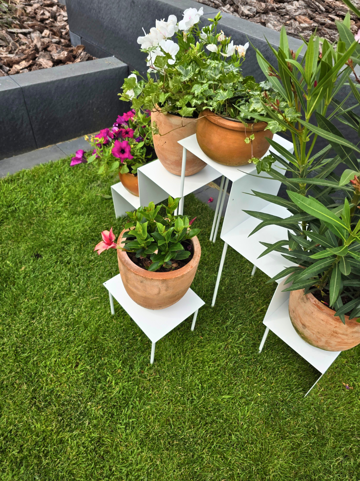 Zdjęcie przedstawia duży kwietnik składający się z trzech różnych wielkości części. Kwietnik jest biały. Stoi na trawie w ogrodzie. Zdjęcie robione z prawej strony kwietnika.