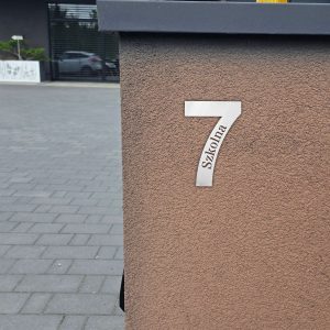 Wizualizacja przedstawia numer na dom w postaci cyfry, w którą wpisana jest nazwa ulicy. Numer zawieszony jest na bramie wjazdowej.