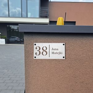 Wizualizacja przedstawia dużą tabliczkę adresową wykonaną z aluminium. Tabliczka ma wycinany laserowo numer, który oddzielony jest pionowym wycięciem od nazwy ulicy. Powieszony jest na bramie wjazdowej.
