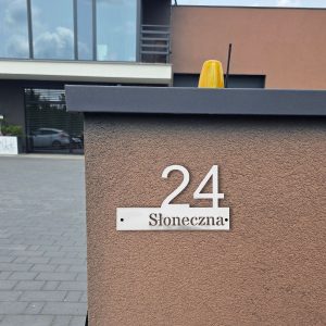 Wizualizacja tabliczki adresowej wykonanej z aluminium. Większą częścią tabliczki jest cyfra 24, do której dołączony jest prostokąt z nazwą ulicy. Tabliczka powieszona na bramie wjazdowej.