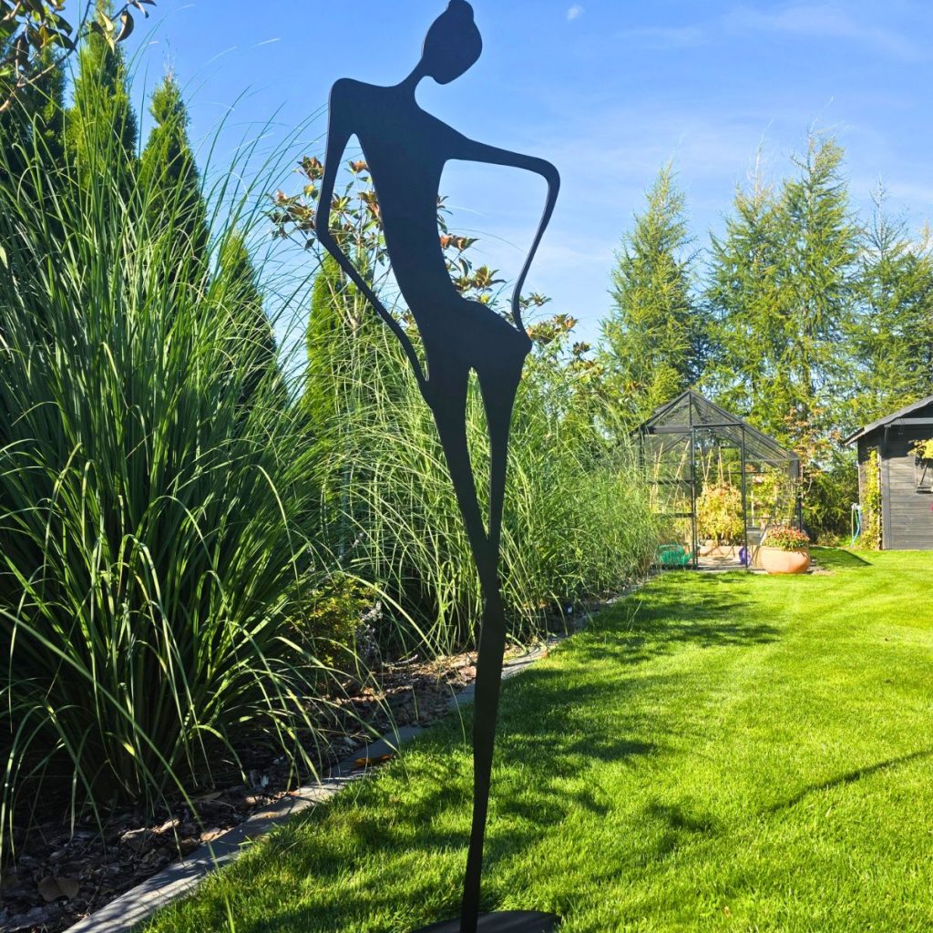 Zdjęcie przedstawia rzeźbę kobiety, która imponuje wysokością. Pozuje na tle ogrodu. Abstrakcyjne kształty dają cały efekt tej nowoczesnej rzeźbie.