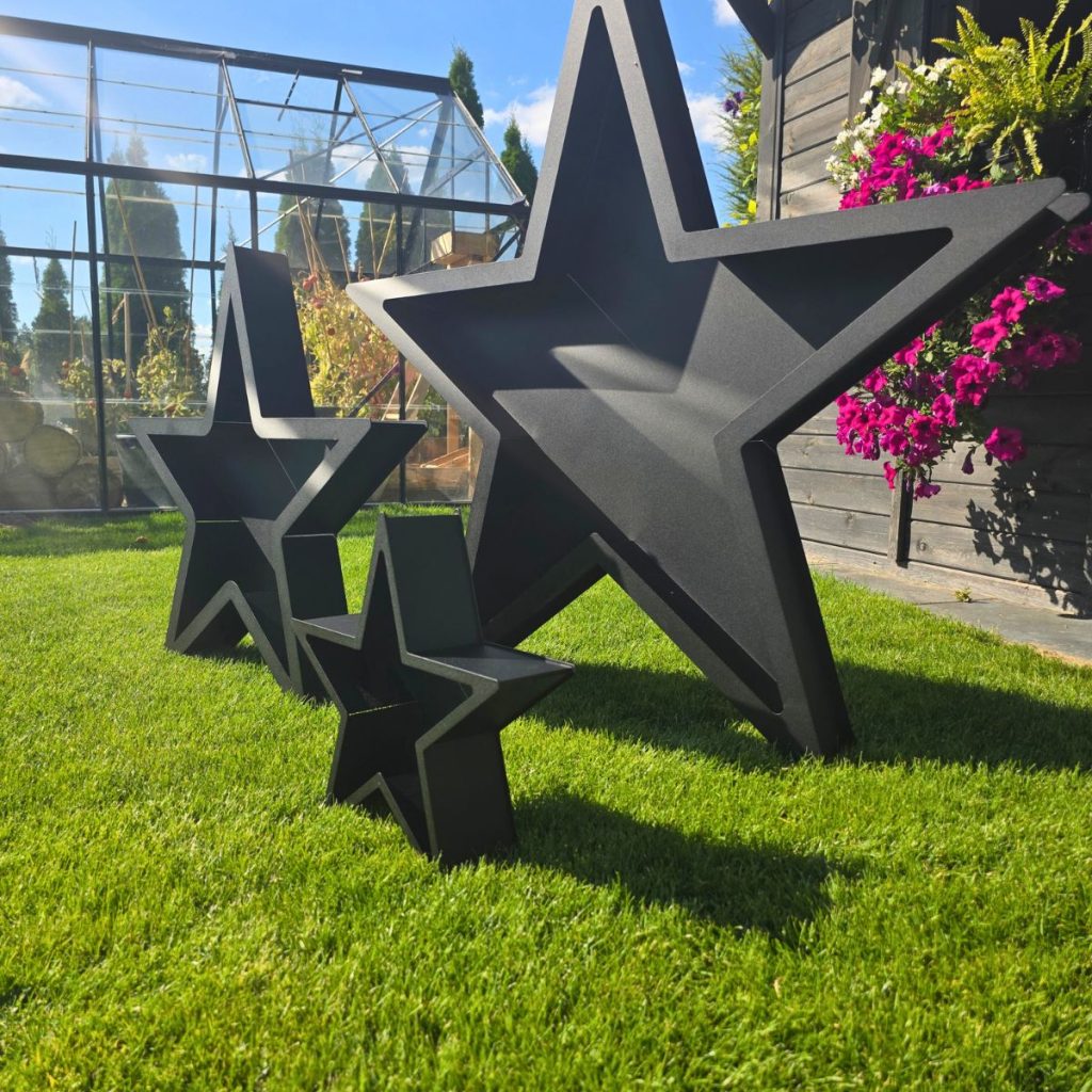 Zdjęcie przedstawia zestaw trzech gwiazd - dekoracji bożonarodzeniowych w różnych rozmiarach. Stoją na zielonej trawie i na tle szklarni.