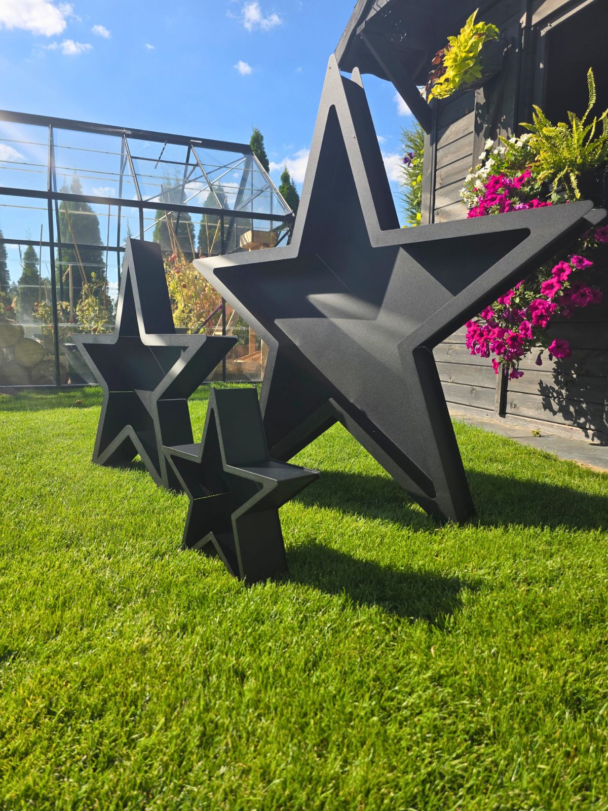 Zdjęcie przedstawia zestaw trzech gwiazd - dekoracji bożonarodzeniowych w różnych rozmiarach. Stoją na zielonej trawie i na tle szklarni.