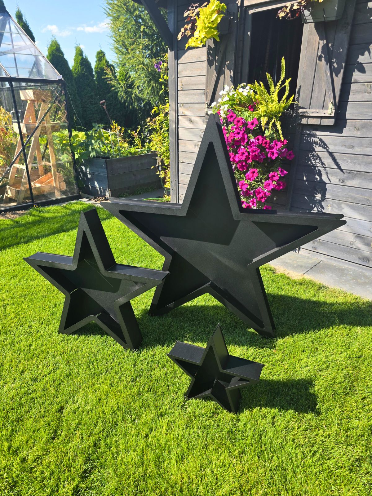Zdjęcie przedstawia zestaw trzech gwiazd - dekoracji bożonarodzeniowych w różnych rozmiarach. Stoją na zielonej trawie i na tle szklarni.