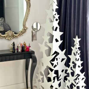 Dwie białe, ażurowe choinki z wyciętych gwiazdek stoją obok ciemnych zasłon w eleganckim salonie; po lewej czarna toaletka z perfumami i ozdobne lustro.