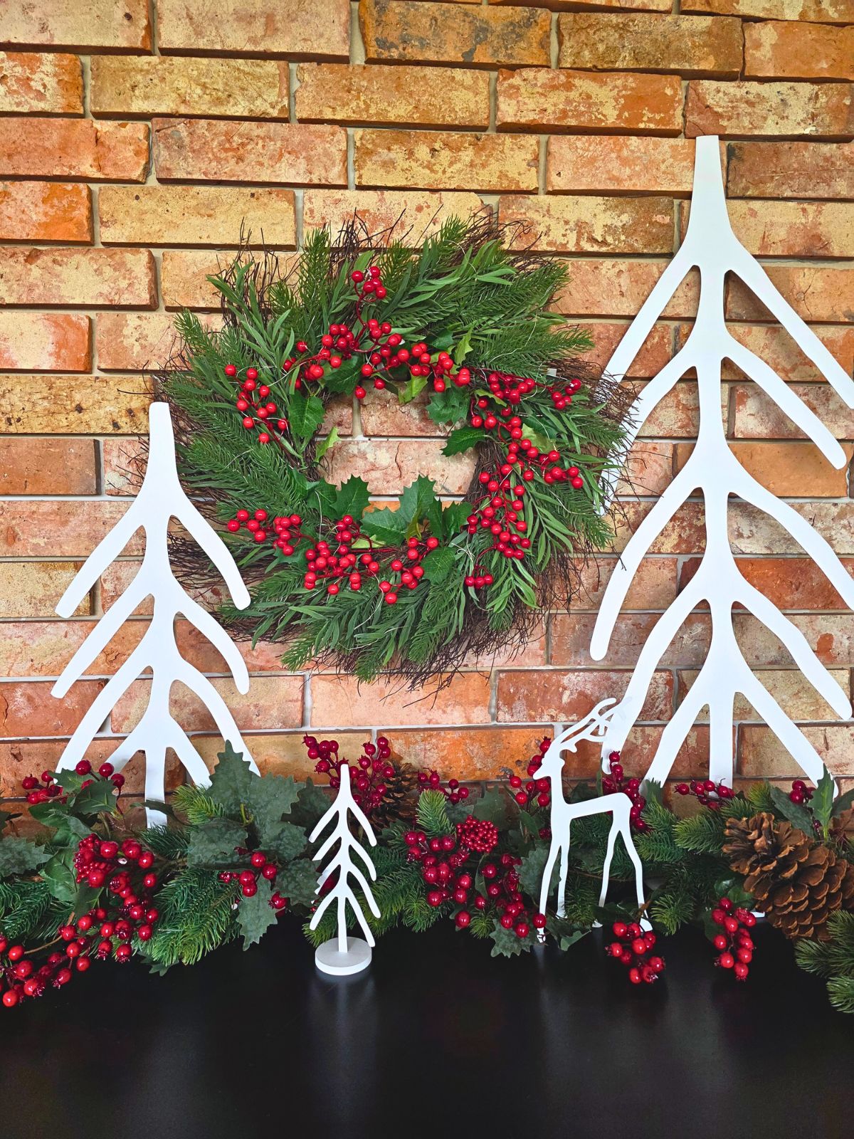 Zdjęcie przedstawia kilka choinek bożonarodzeniowych oraz renifera. Stoją na tle ceglanej ściany w otoczeniu łańcucha iglastego oraz czerwonych owoców jarzębiny.