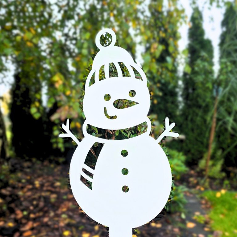 Zdjęcie przedstawia wbitego w ziemię bałwanka - ozdobę metalową o takim wzorze. Bałwanek stoi na tle drzew - tuje i brzoza.