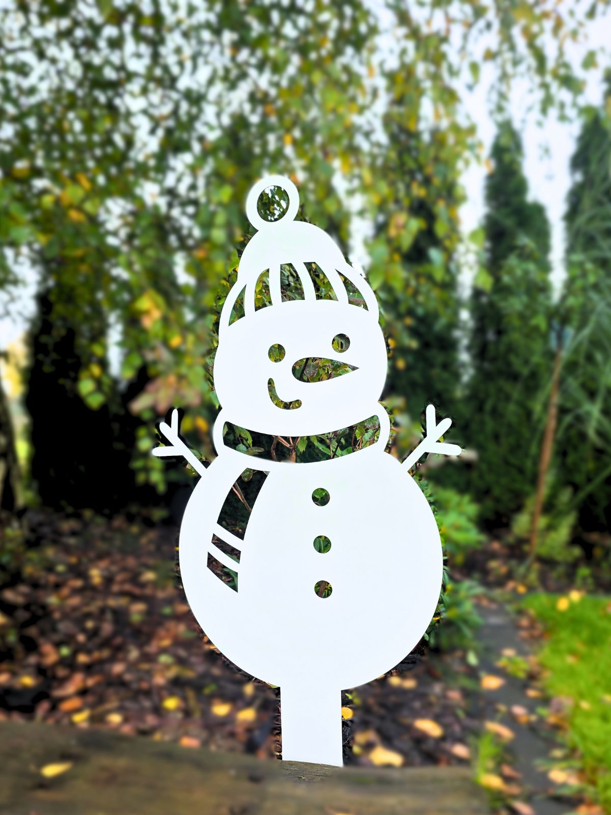 Zdjęcie przedstawia wbitego w ziemię bałwanka - ozdobę metalową o takim wzorze. Bałwanek stoi na tle drzew - tuje i brzoza.