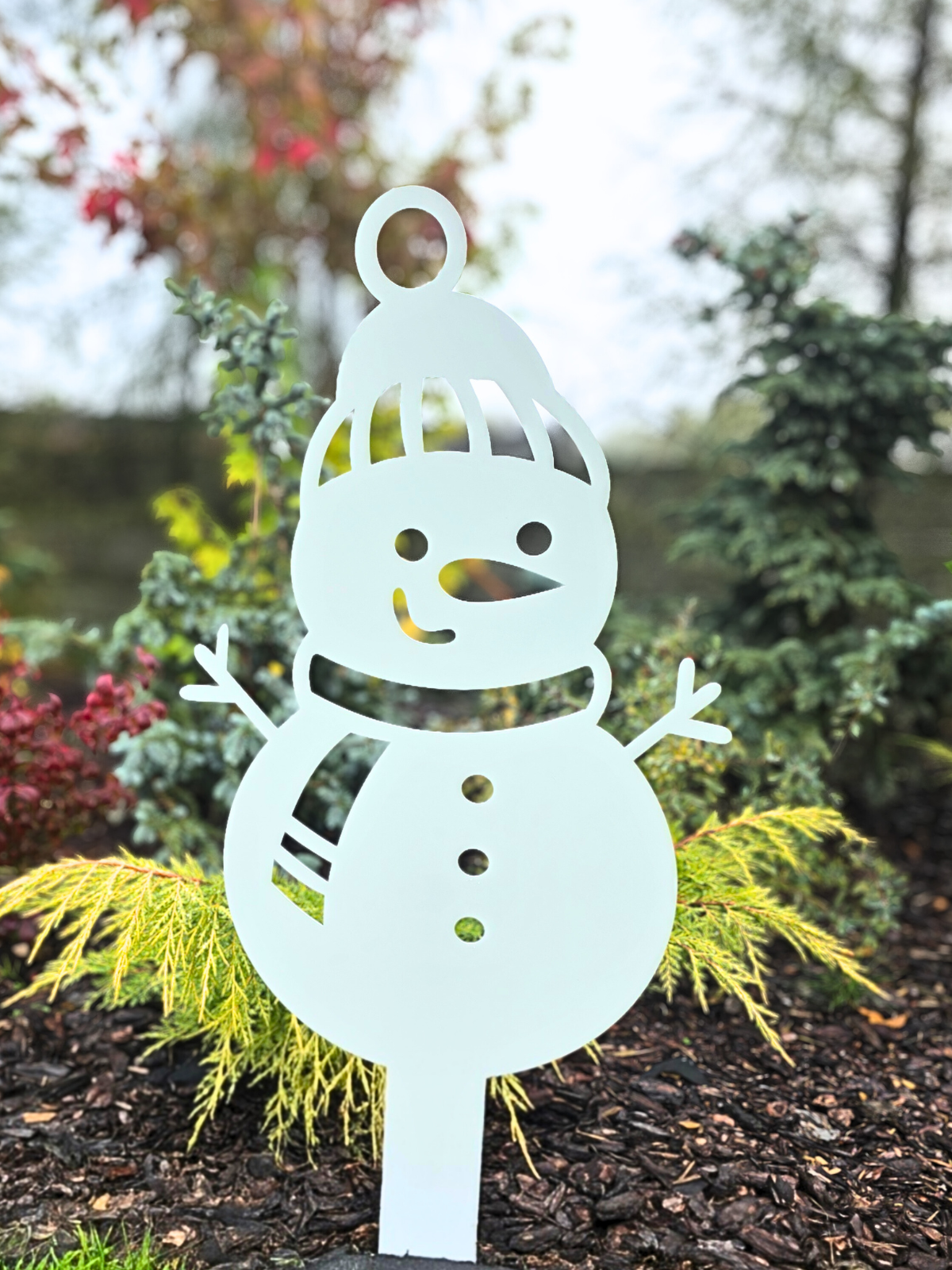 Zdjęcie przedstawia faktyczny produkt- wbijany bałwan. Ozdoba świąteczna wbita jest w ziemię pokrytą korą. W tle widać roślinność ogrodową taką jak drzewa iglaste, krzaczki.