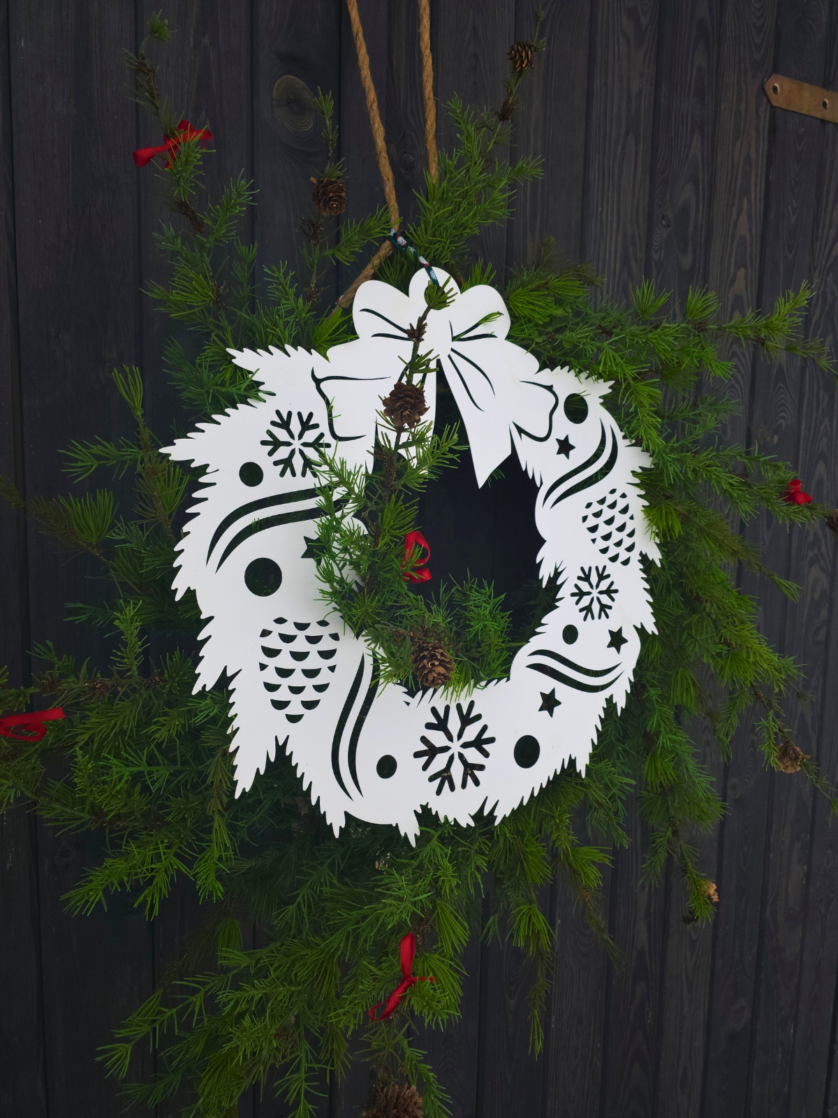 Papierowy wieniec z wyciętymi śnieżynkami i szyszkami, zawieszony wśród świeżych gałązek iglastych.