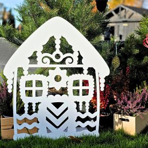 Zdjęcie przedstawia biały domek z piernika wykonany z metalu. Domek stoi na tle choinek, wrzosów.