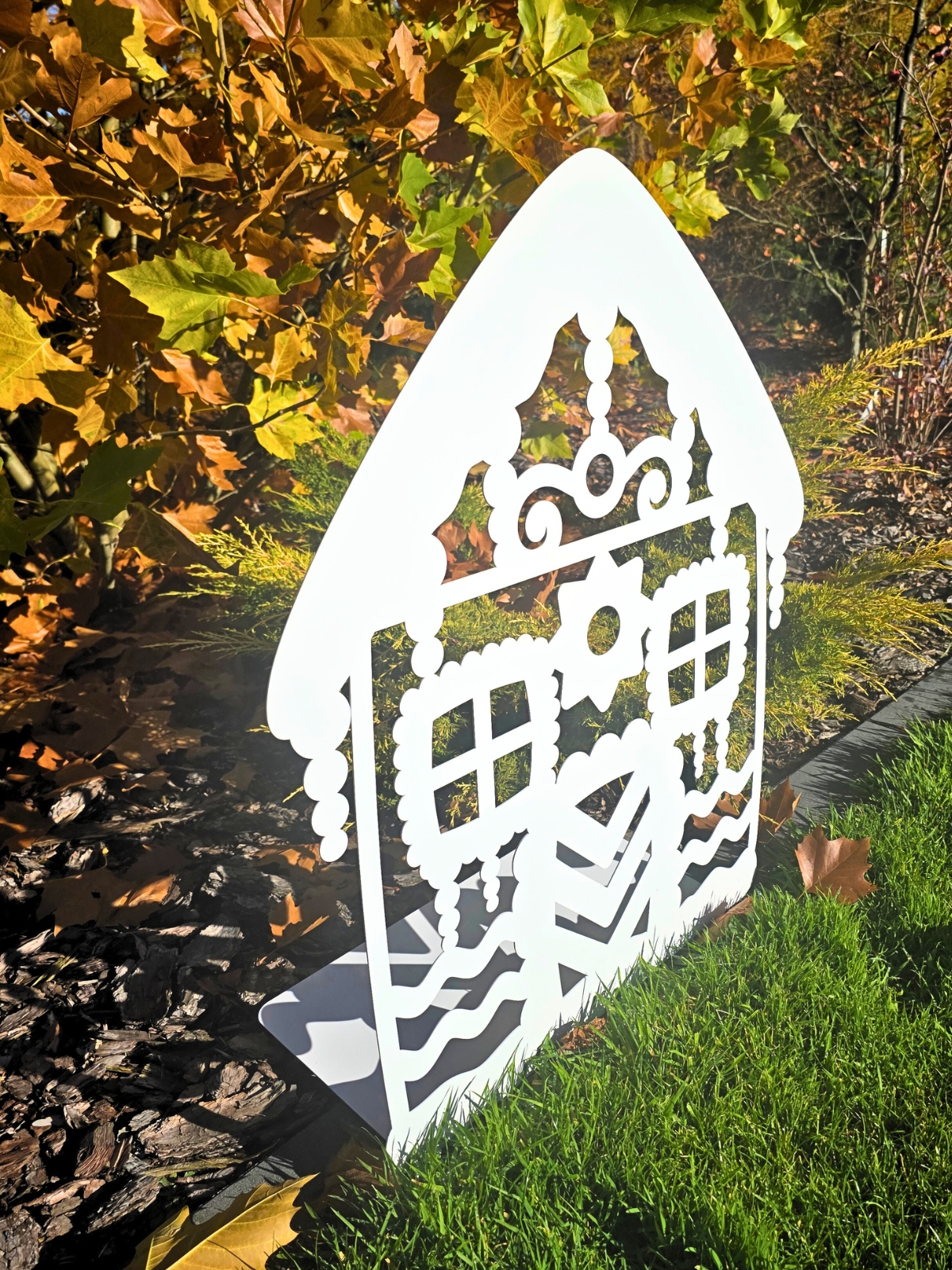 Zdjęcie przedstawia biały domek z piernika wykonany z metalu. Domek stoi na tle żółtych liści, krzewów. Domek stoi bokiem, widać zaginaną podstawkę, która utrzymuje domek.