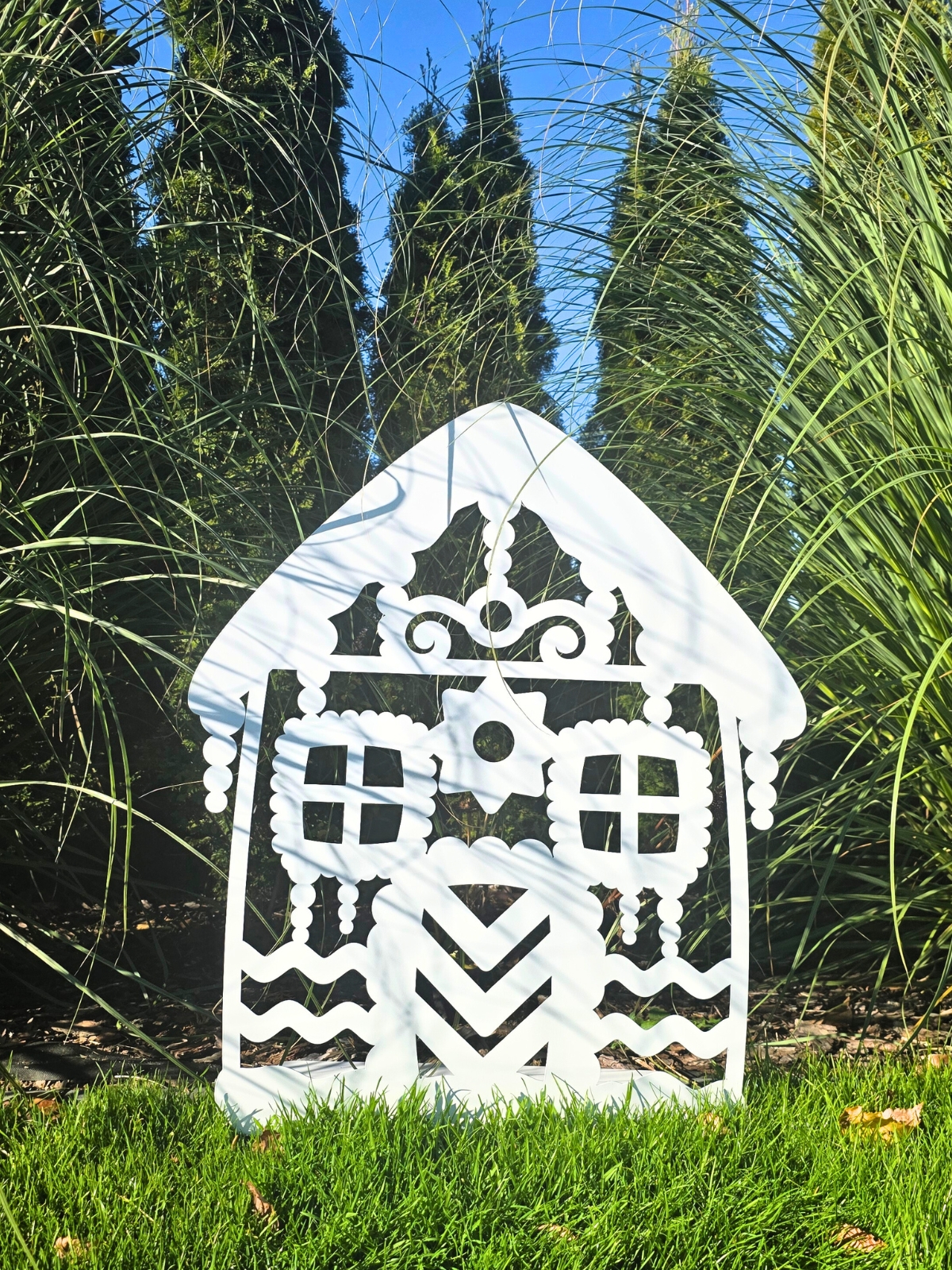 Zdjęcie przedstawia biały domek z piernika wykonany z metalu. Domek stoi na tle drzewek.