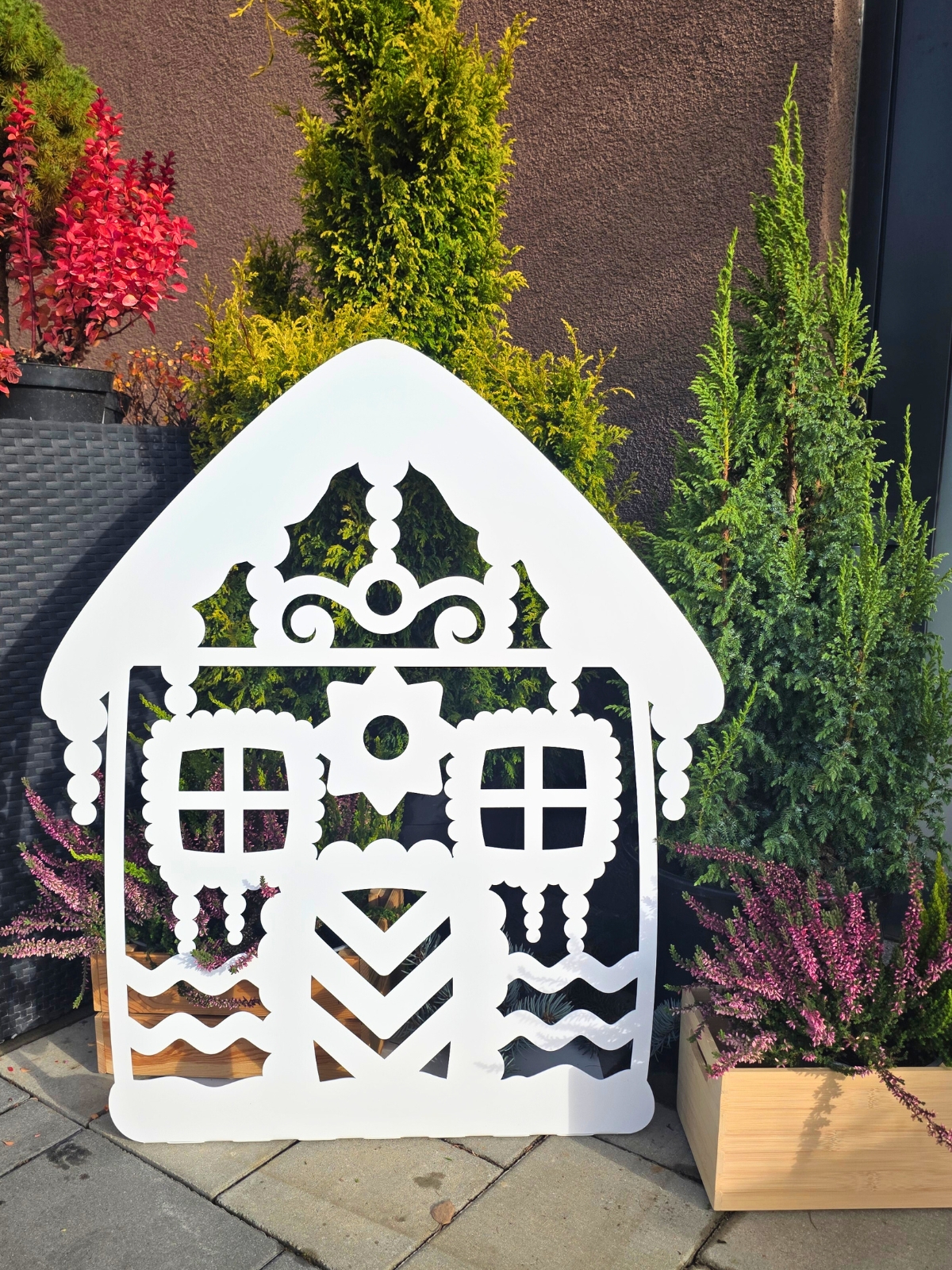 Zdjęcie przedstawia biały domek z piernika wykonany z metalu. Domek stoi na tle choinek, wrzosów przed wejściem do domu.