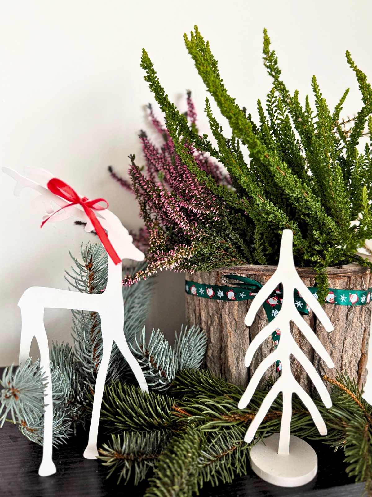 Kompozycja świąteczna na półce: białe figurki renifera i choinki wśród zielonych gałązek, w tle donica z wrzosami ozdobiona wstążką.