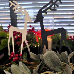 Dwie dekoracyjne figurki reniferów, jasna i ciemna, stoją naprzeciw siebie wśród zielonych gałązek, czerwonych jagód i świeczki w tle.