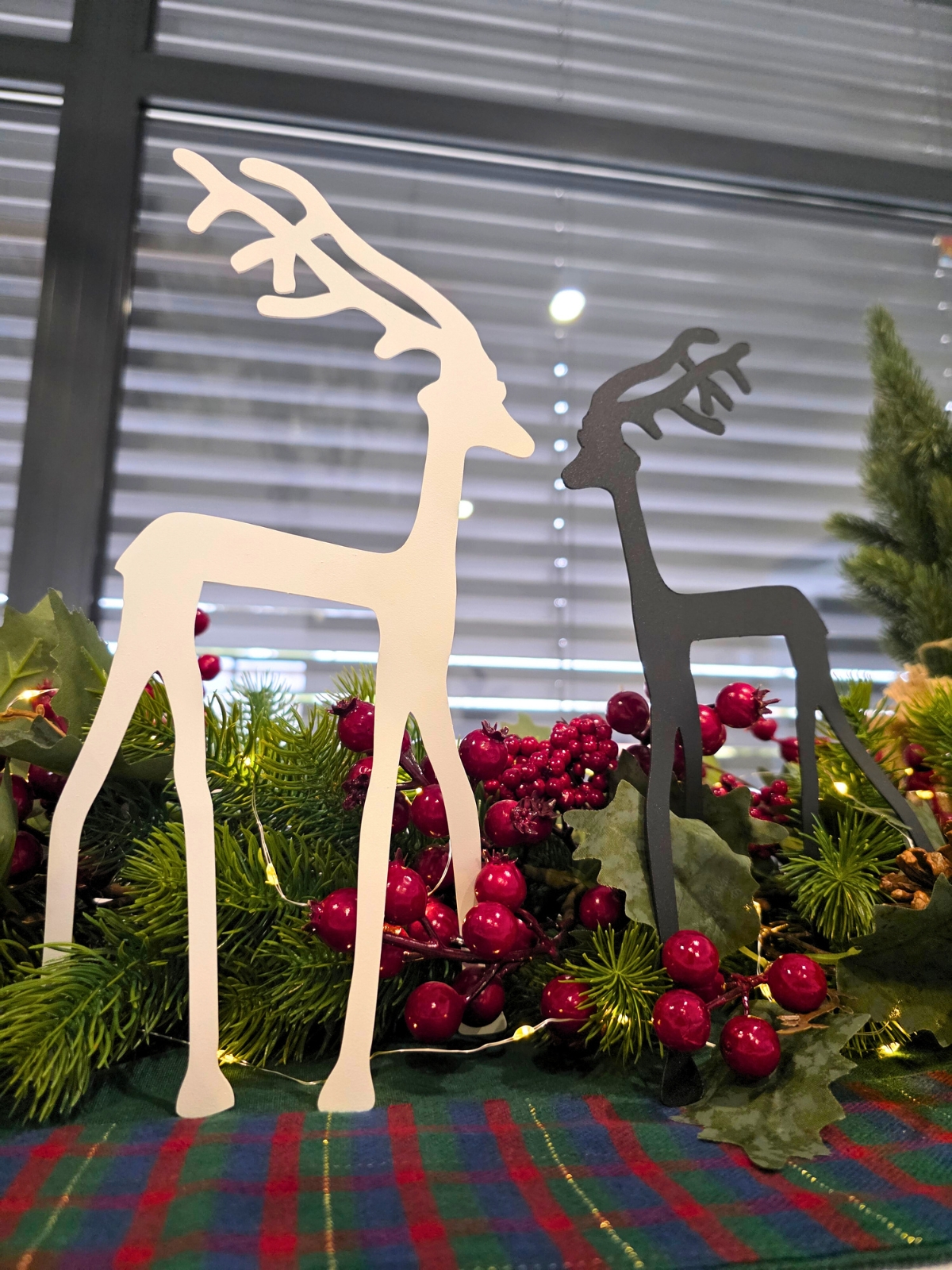Świąteczna aranżacja: jasny i czarny renifer z wyciętego metalu, otoczone gałązkami, szyszkami i czerwonymi jagodami na kraciastej serwecie.
