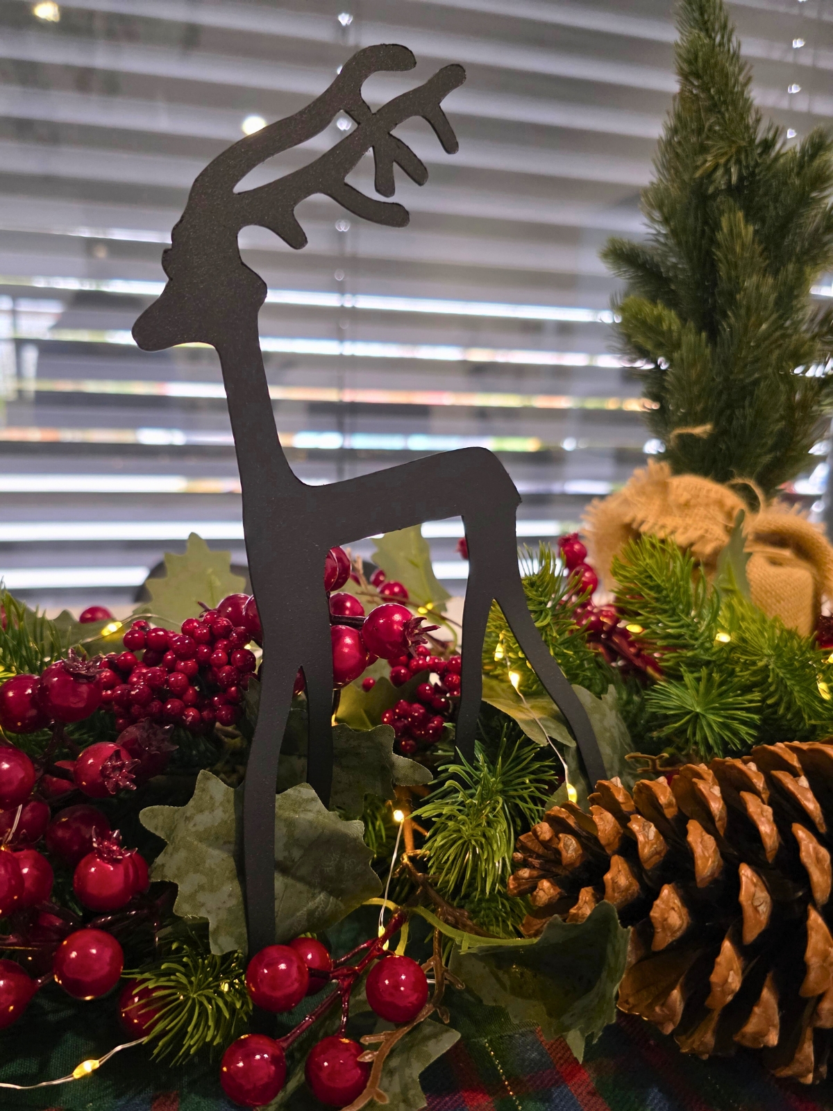 Czarna sylwetka renifera w dekoracji bożonarodzeniowej z zielonymi igłami, czerwonymi owocami i lampkami.