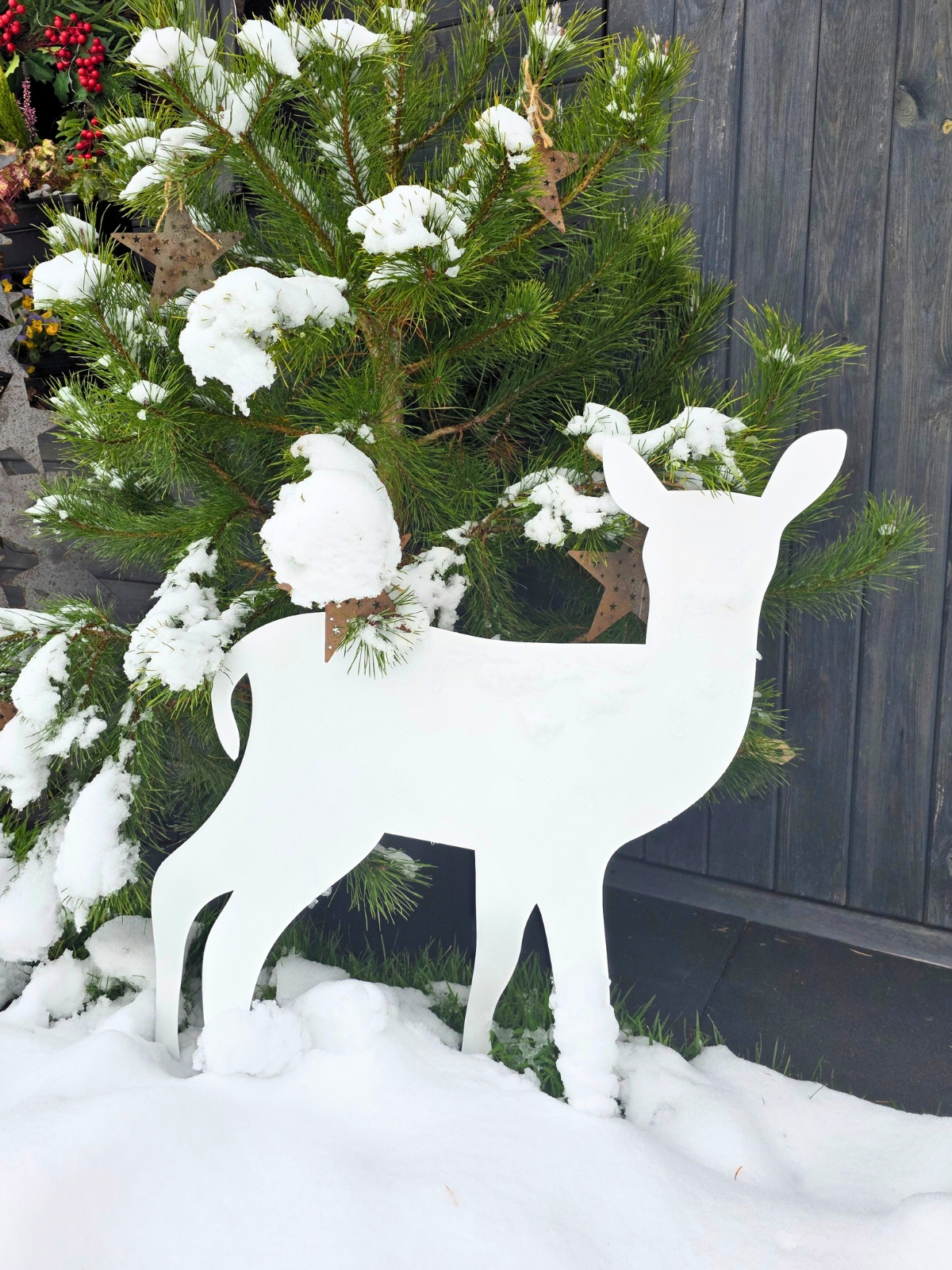 Biała, płaska, metalowa sylwetka sarenki stoi w śniegu przed zielonym, ośnieżonym krzewem na tle ciemnej, drewnianej ściany