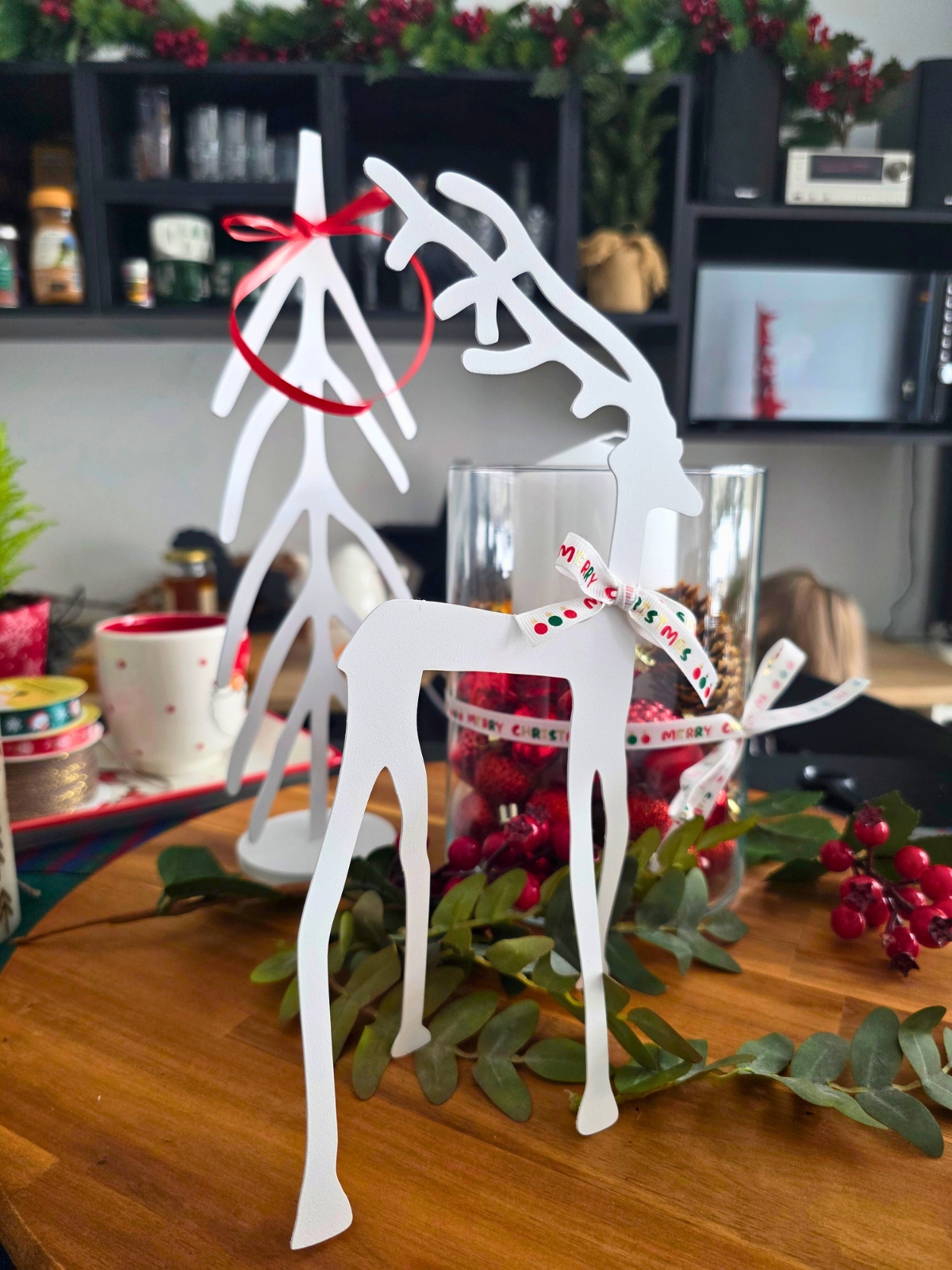 Białe dekoracyjne renifery stojące na drewnianym blacie otoczone zielonymi gałązkami i czerwonymi świątecznymi ozdobami.