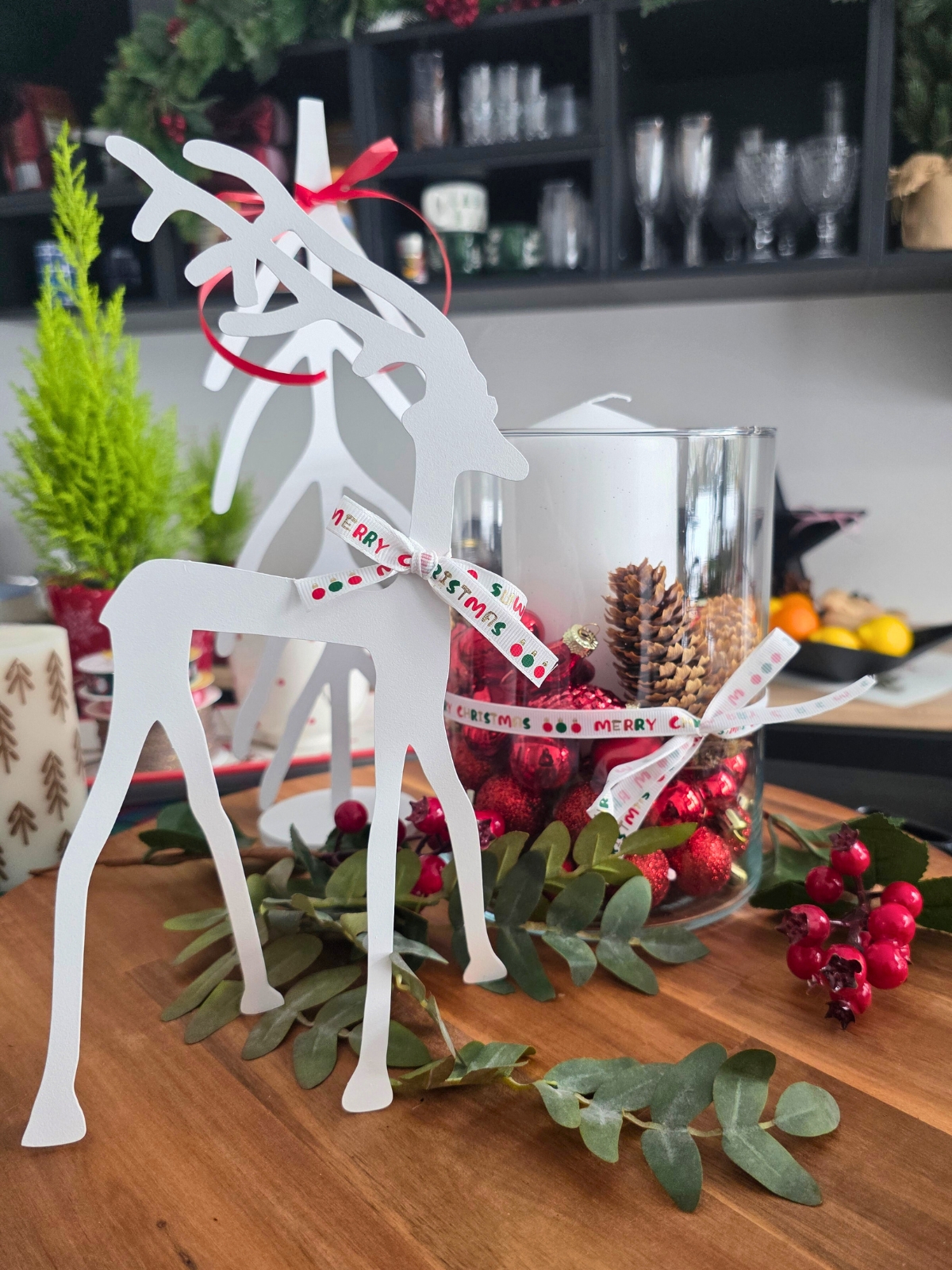 Białe dekoracyjne renifery stojące na drewnianym blacie otoczone zielonymi gałązkami i czerwonymi świątecznymi ozdobami.