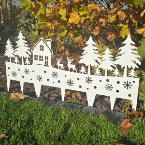 Biała dekoracja ogrodowa przedstawiająca domek, drzewa i dwa jelenie na tle jesiennych liści.