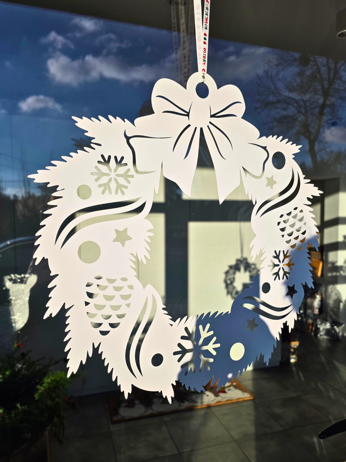 Papierowy, biały wieniec świąteczny z wyciętymi wzorami (śnieżynki, bombki, gwiazdki) zawieszony na wstążce na szybie, w tle odbicie okna i niebo z chmurami.