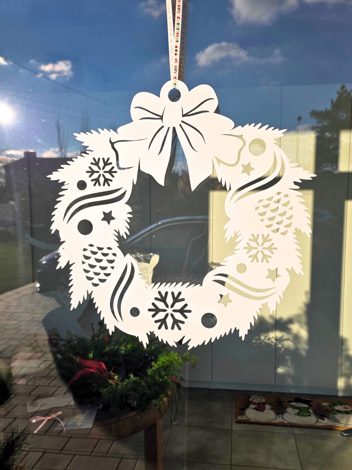 Papierowy, biały wieniec świąteczny z wyciętymi wzorami (śnieżynki, bombki, gwiazdki) zawieszony na wstążce na szybie, w tle odbicie okna i niebo z chmurami.