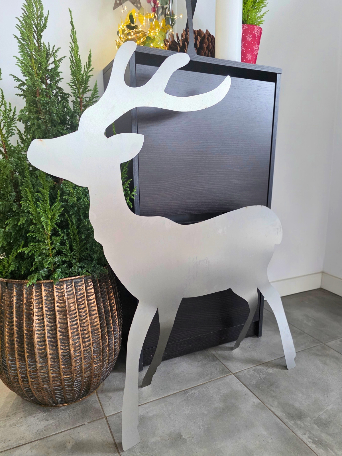 Metalowy renifer stojący na podłodze obok zielonej rośliny w donicy.
