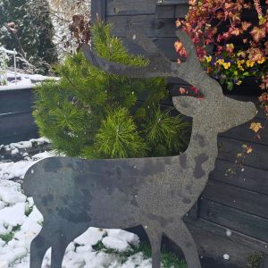 Metalowy kształt jelenia stojący na śniegu obok drewnianej ściany i zimowych roślin.