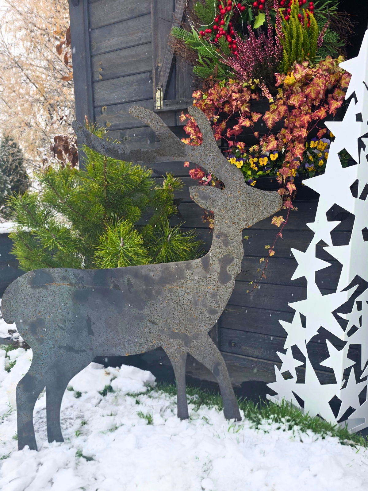 Metalowy kształt jelenia stojący na śniegu obok drewnianej ściany i zimowych roślin.