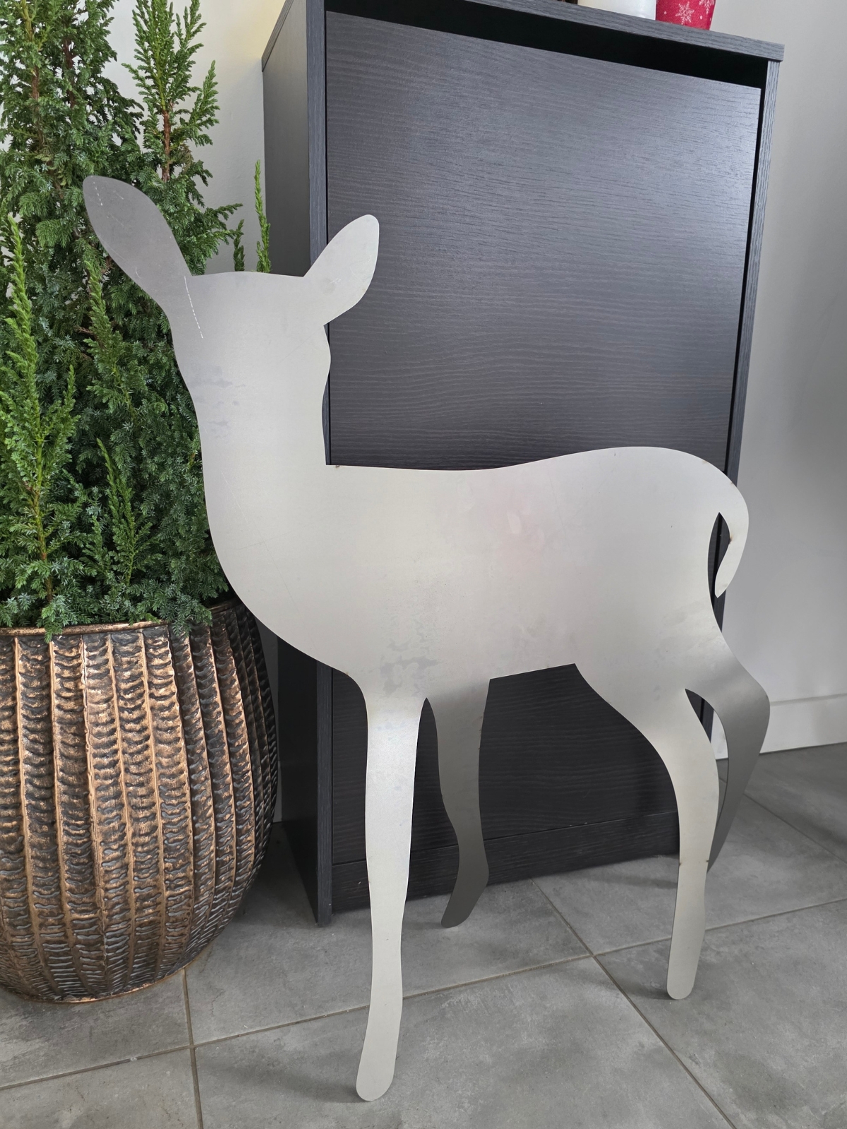 Metalowy dekor w kształcie młodej sarny stojącego na podłodze obok wysokiej rośliny.