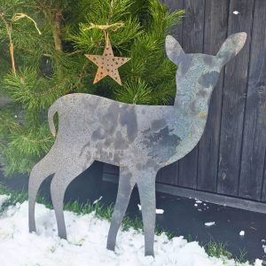 Metalowa sylwetka sarny stojąca w śniegu obok zielonego drzewka z gwiazdkami.
