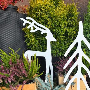 Minimalistyczny biały renifer i choinka stoją obok drewnianych skrzynek z roślinami i wysokiej donicy, tło z gęstych drzew iglastych