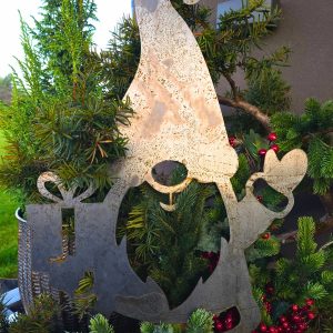 Metalowa dekoracja świąteczna w kształcie skrzata z długą czapką, wśród zielonych iglaków i czerwonych jagód.