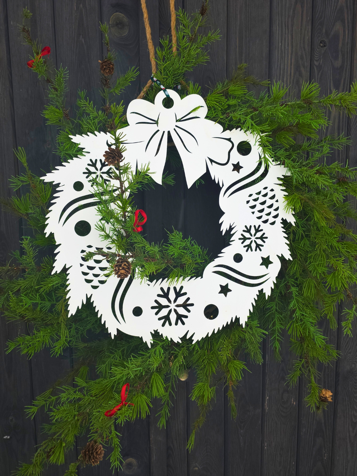 Papierowy wieniec z wyciętymi śnieżynkami i szyszkami, zawieszony wśród świeżych gałązek iglastych.