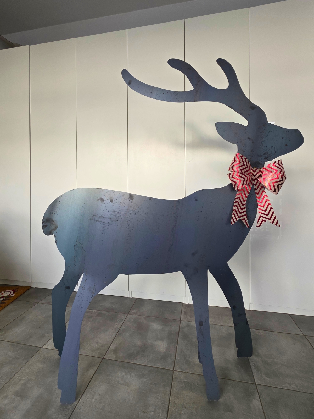 Płaska figurka metalowego renifera który ma 165 cm wysokości z ozdobną kokardą, ustawiona na tle jasnej ściany.