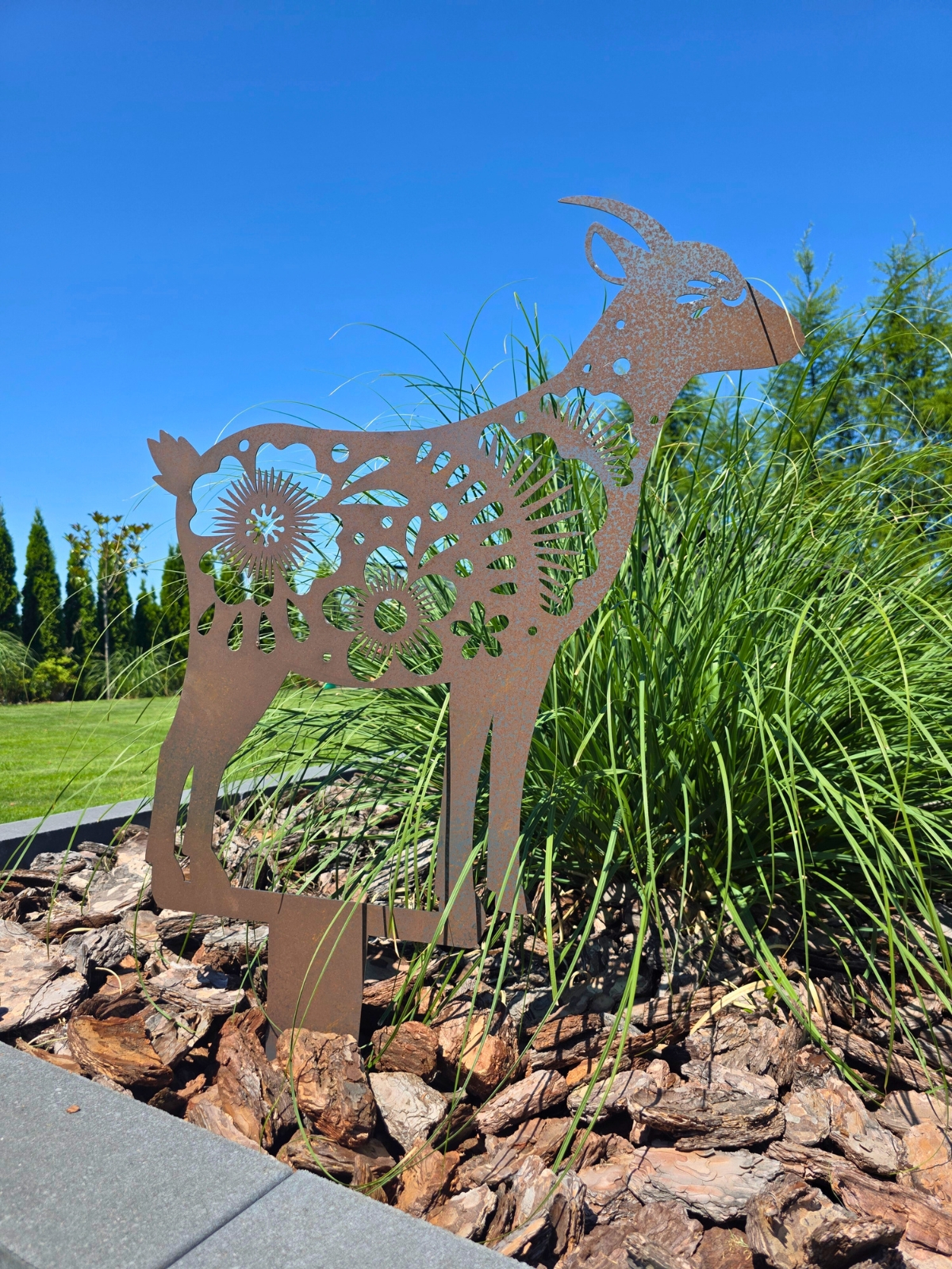 Metalowa dekoracja ogrodowa w kształcie kozy z ażurowymi kwiatowymi wzorami stojąca na tle zielonej trawy.