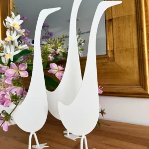 Trzy białe dekoracyjne figurki ptaków stojące na drewnianej powierzchni obok różowych i białych kwiatów.