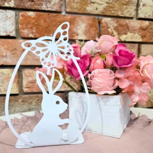 Ozdoba w kształcie królika z motylem obok bukietu różowych róż ustawionych w białym pojemniku.