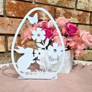 Biała, ażurowa dekoracja w kształcie jajka z króliczkiem, kwiatami i motylem, stojąca przed bukietem różowych i czerwonych kwiatów.