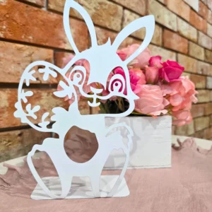 Ozdobny biały króliczek z ażurowymi detalami stojący przed bukietem różowych kwiatów na tle ceglanej ściany.