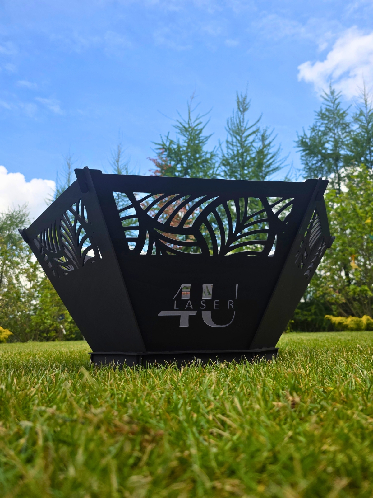 Czarny, metalowy palenisko ogrodowe stojące na zielonym trawniku, na tle drzew.