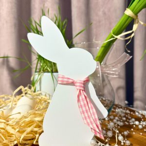 Biała figurka zająca z różową wstążką stojąca na drewnianym blacie, otoczona wielkanocnymi dekoracjami.