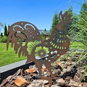 Ażurowy kogut z metalu ustawiony w ogrodowej rabacie wśród kory drzewnej.