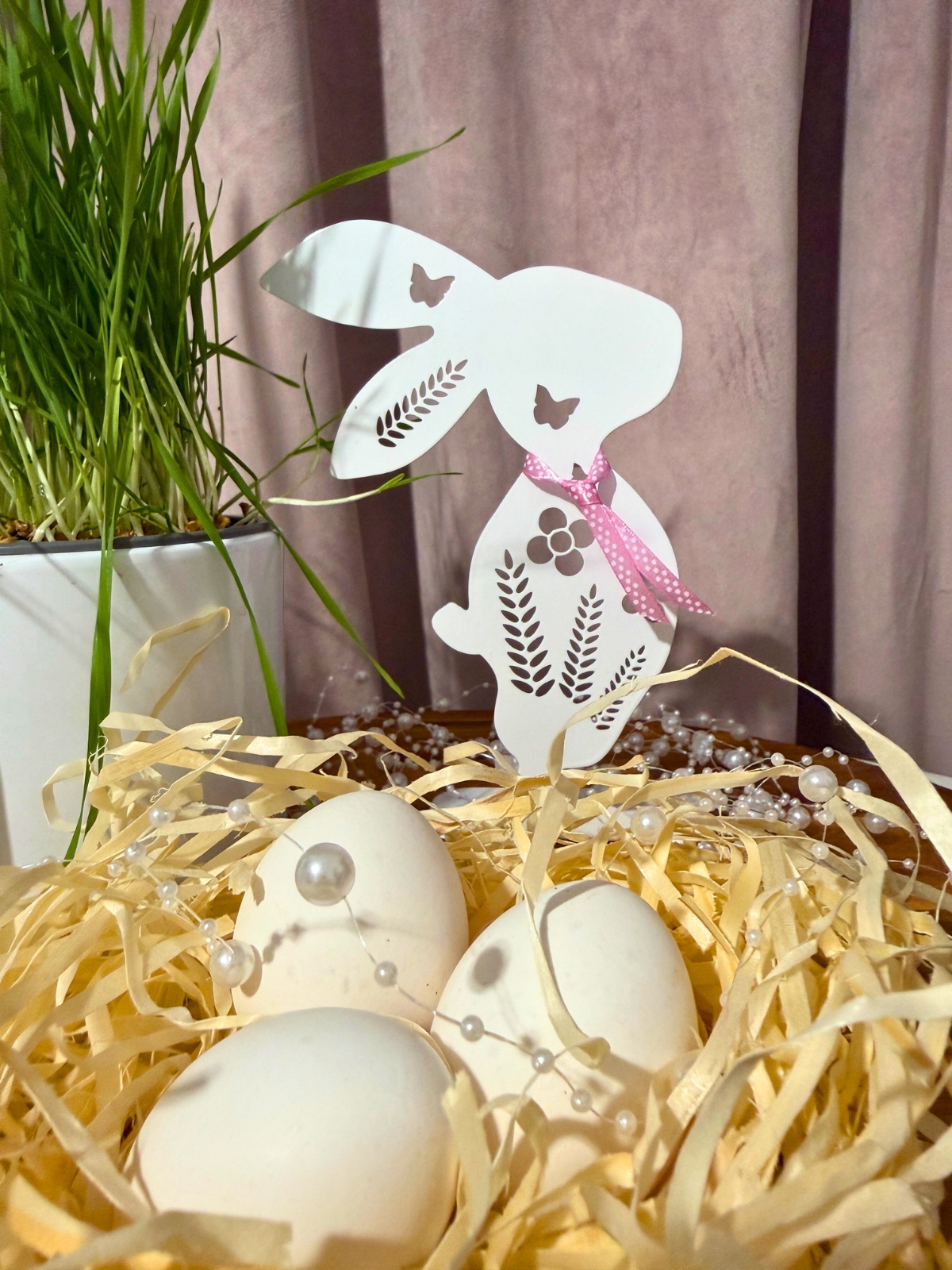 Świąteczna kompozycja z białym króliczkiem, jajkami i trawą w doniczce, ułożona w naturalnym, ciepłym otoczeniu.