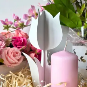 Metalowy biały tulipan obok różowej świecy wśród róż i ozdobnych dodatków.