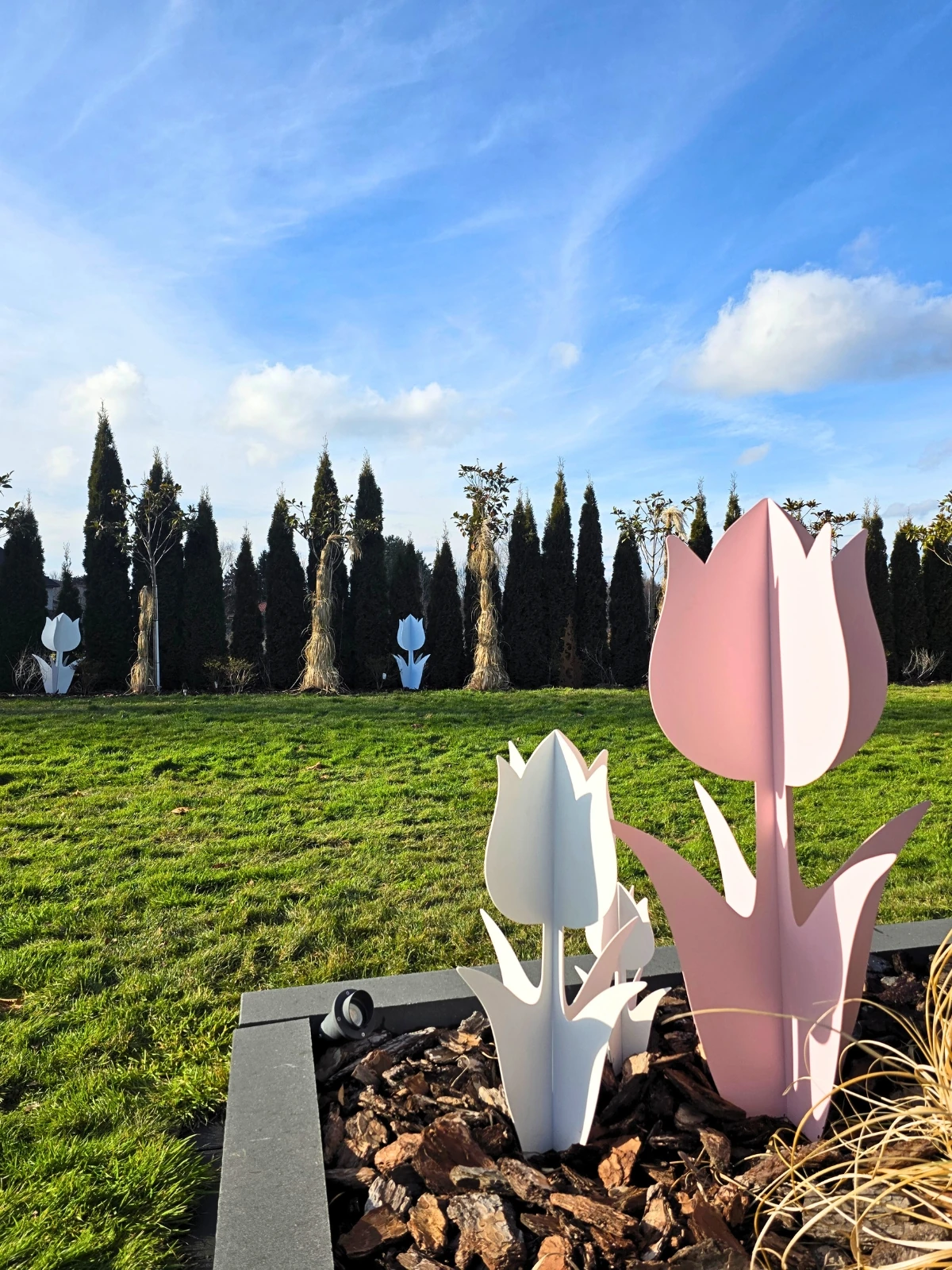 Metalowe tulipany stoją na tle zielonego trawnika i wysokich drzew, oświetlone jasnym, słonecznym niebem.